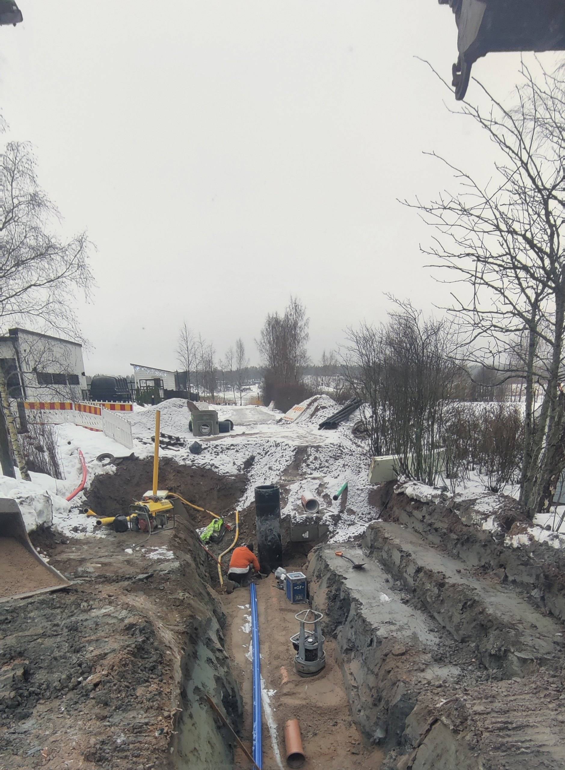 Syvä putkikaivanto talvella, kaivannon pohjalla vesi- ja viemäriputkia ja työntekijä